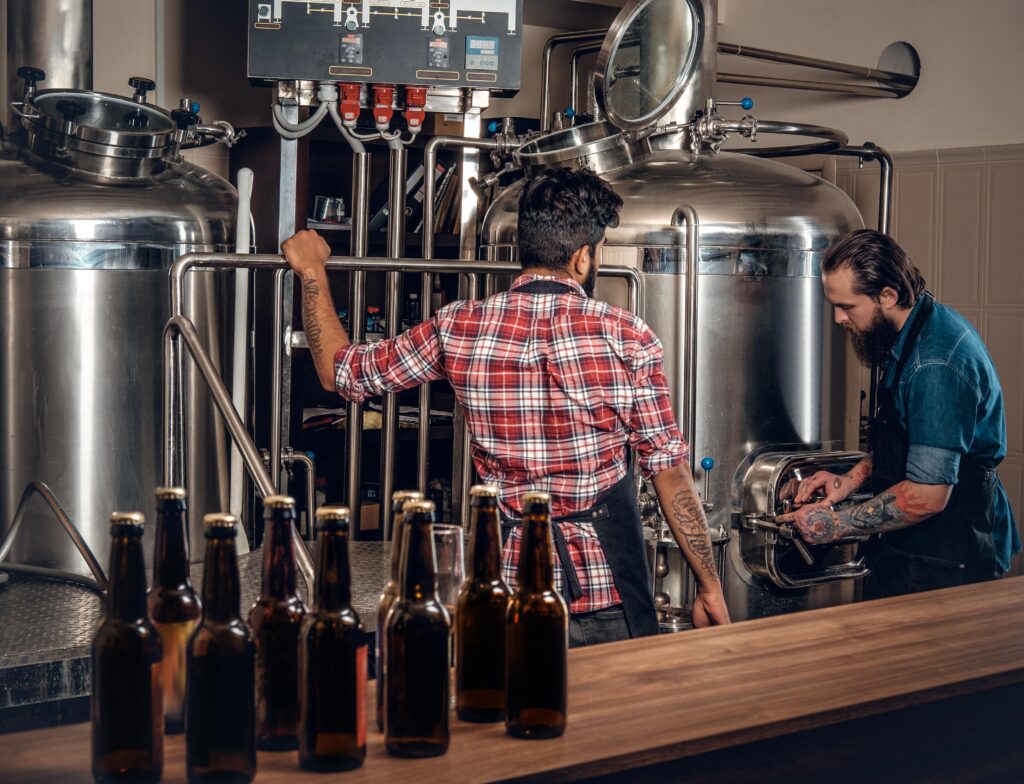 Two brewers working in a craft brewery. One man leans against a stainless steel tank, while the other checks on a bottling line. Bottles of beer are lined up on a counter, ready for distribution.