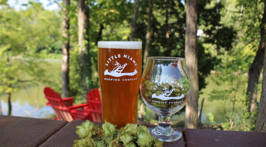 A pint of beer and a glass of beer next to hops, with a scenic view of the Little Miami River in the background.