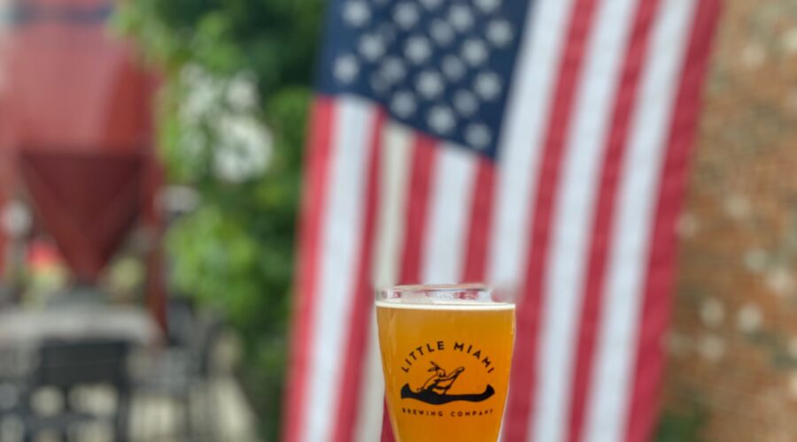 A hand holds a Little Miami Brewing beer glass with an American flag backdrop.