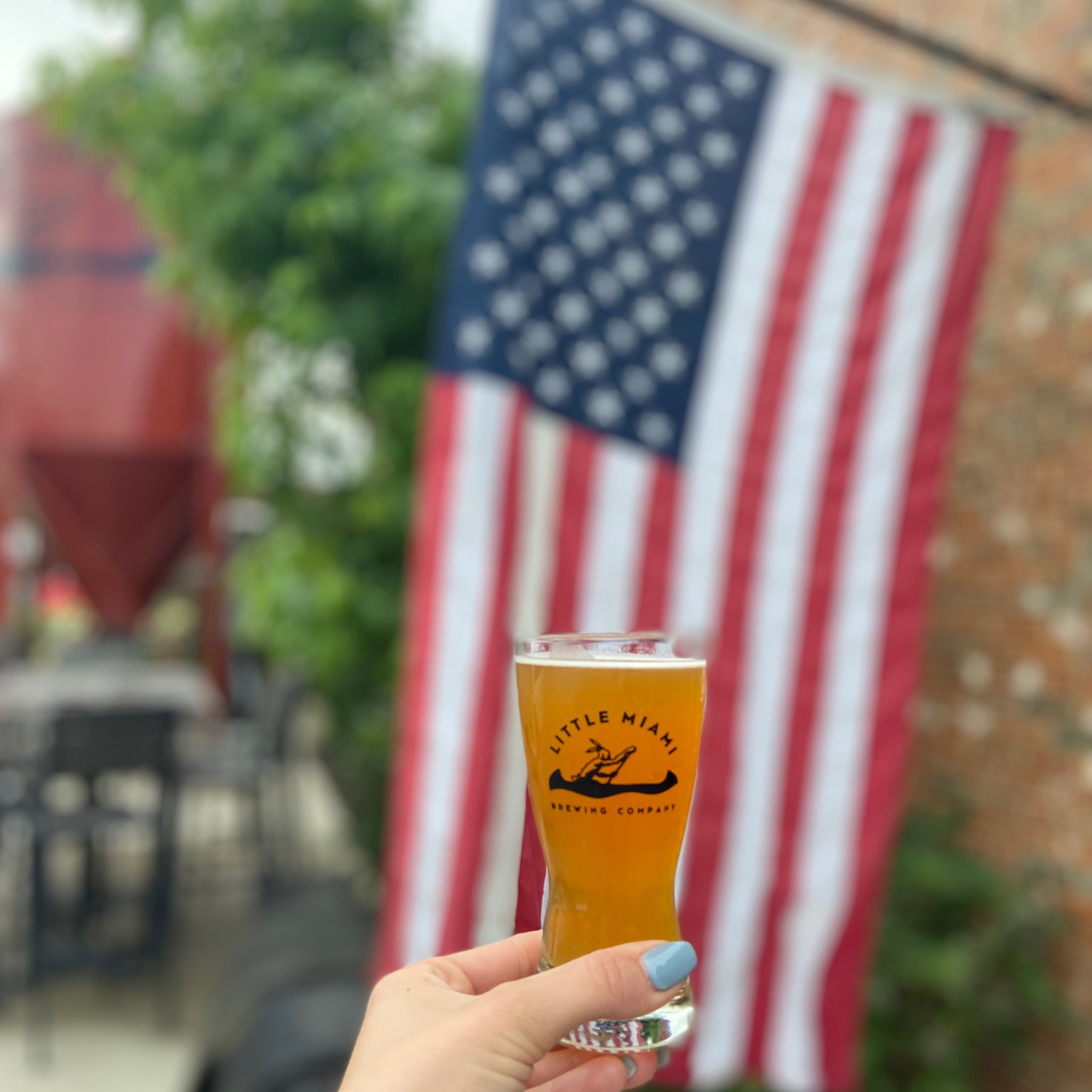 A hand holds a Little Miami Brewing beer glass with an American flag backdrop.