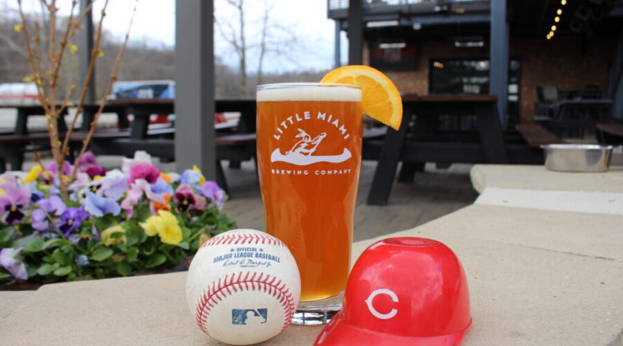 LMBC’s Curveball Beer, a baseball, and a red C-logo helmet on a stone ledge with flowers and outdoor seating.