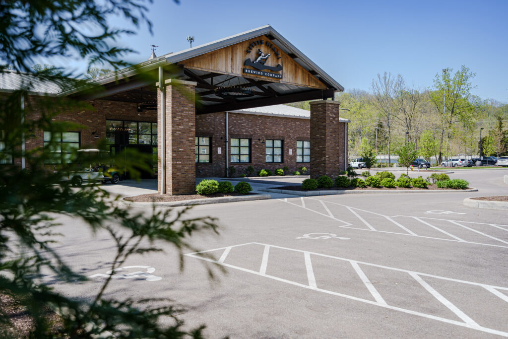 The exterior of Little Miami Brewing Company with its entrance, parking lot, and trees under clear skie.