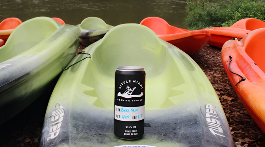 A can of Little Miami Brewing Company beer rests on a green kayak, with other kayaks and the Little Miami River in the background.