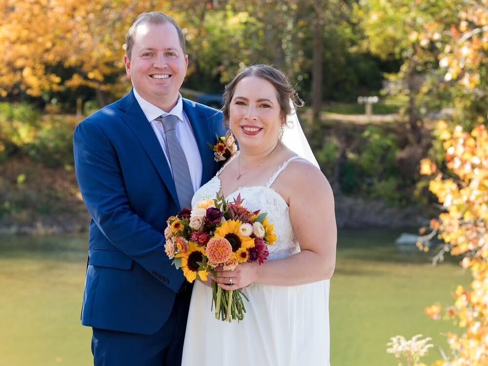 Bride and groom pose in front of the Little Miami River with golden fall foliage during an outdoor wedding ceremony.