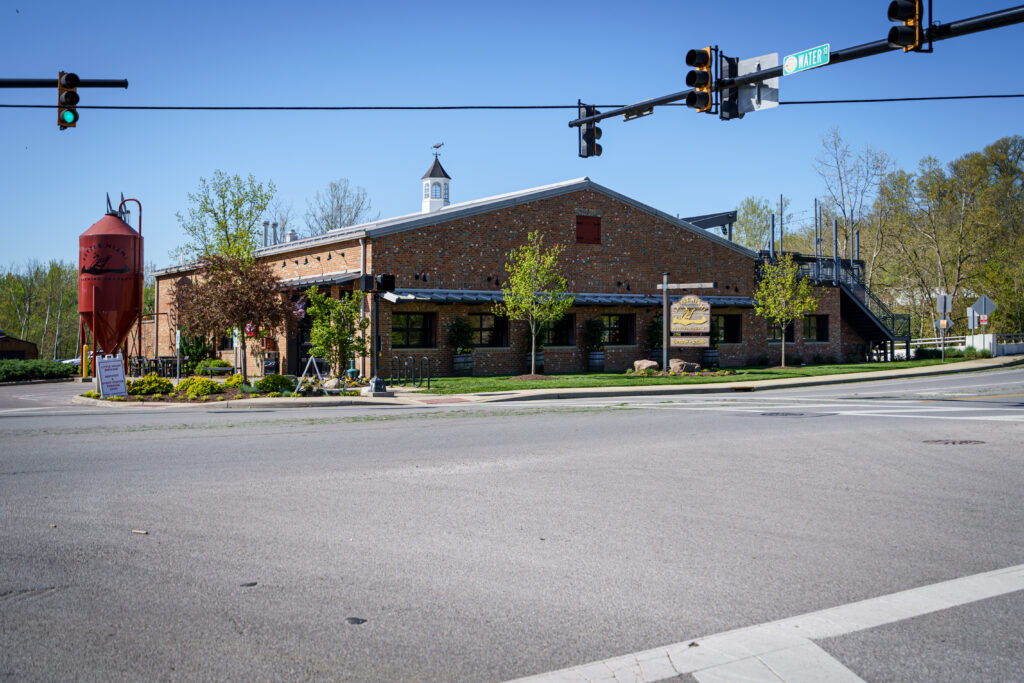 Exterior of Little Miami Brewing Company with its red silo, rustic brick walls, and trees on a sunny day.