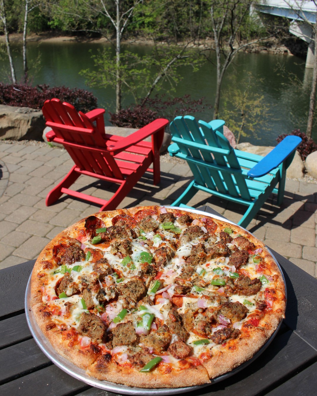 Brick-oven pizza from Little Miami Brewing Company served on a riverside table with red and blue chairs overlooking the Little Miami River.