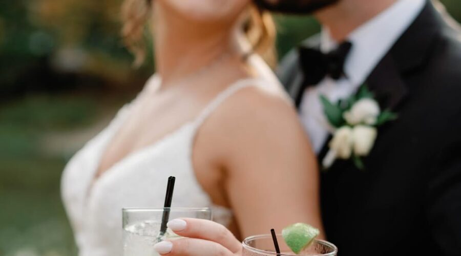 Bride smiles as groom kisses her cheek, both holding cocktails at Little Miami Brewing Company by the riverside.