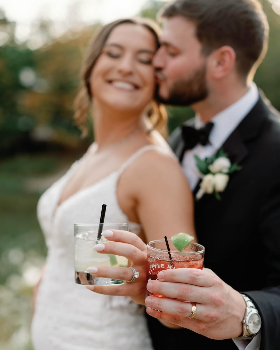 Bride smiles as groom kisses her cheek, both holding cocktails at Little Miami Brewing Company by the riverside.
