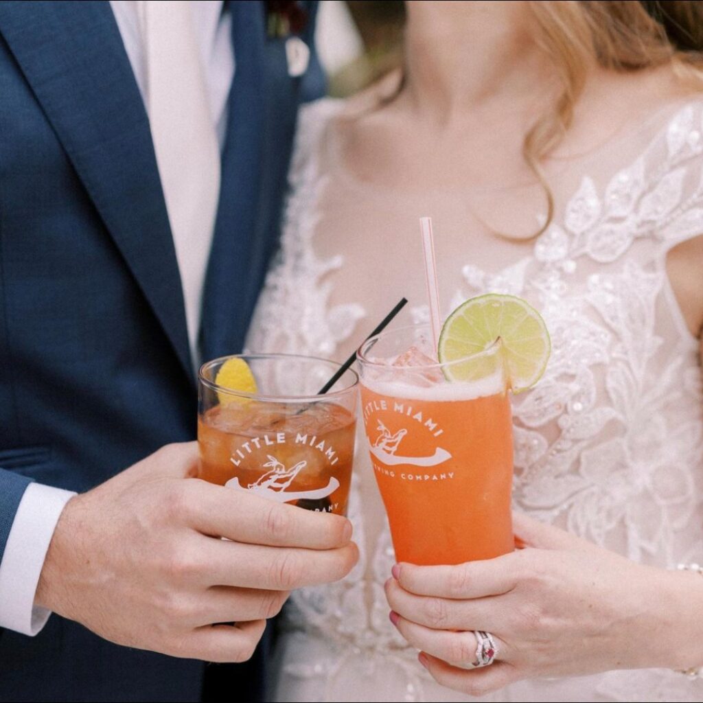 Bride in a white lace dress and groom in a blue suit clinking beer glasses at Little Miami Brewing Company.