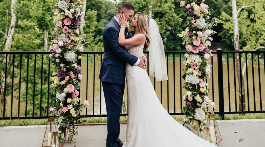 A couple standing beneath a blooming floral arch at Little Miami Brewing Company, with the Little Miami River in the background.