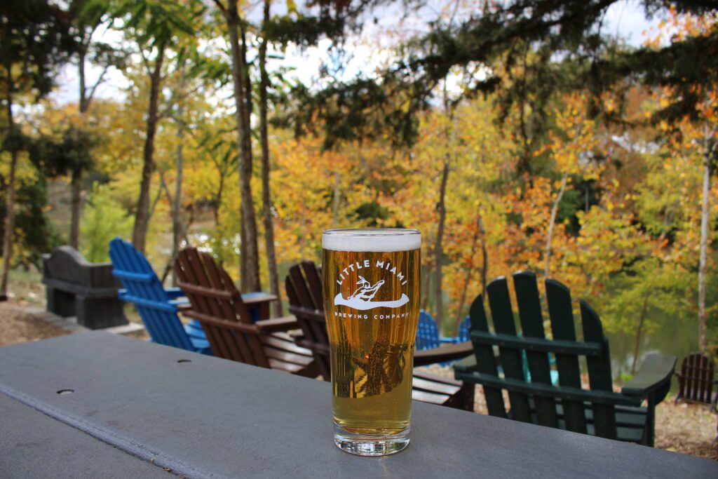 A pint of Bike Path Pilsner from Little Miami Brewing Company, with outdoor chairs and colorful autumn trees in the background.