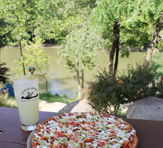 A loaded pizza and a pint from Little Miami Brewing Company on a wooden table in front of a lush riverside.