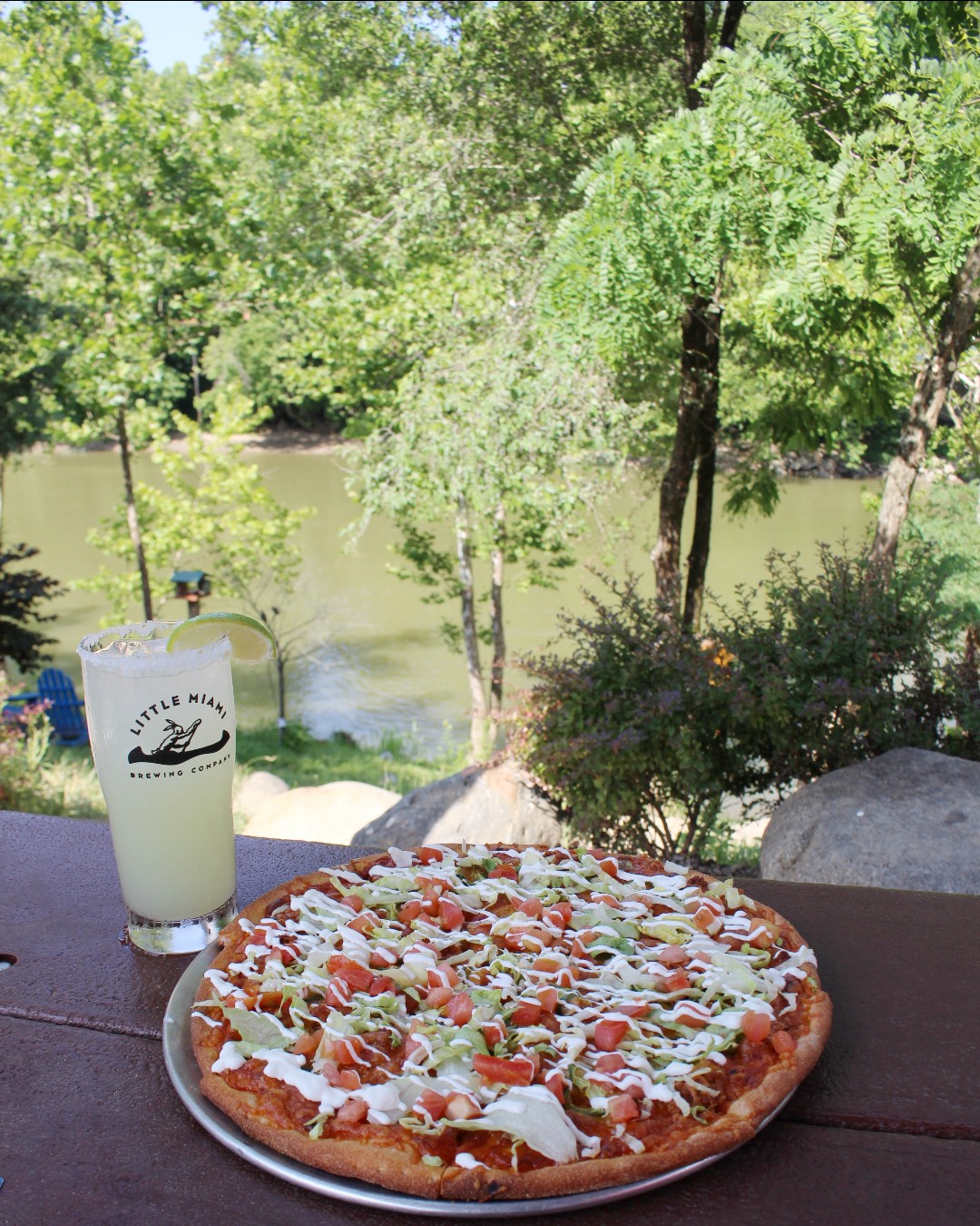 A pizza and margarita cocktail on a rustic wooden table overlooking the Little Miami River at Little Miami Brewing Company.