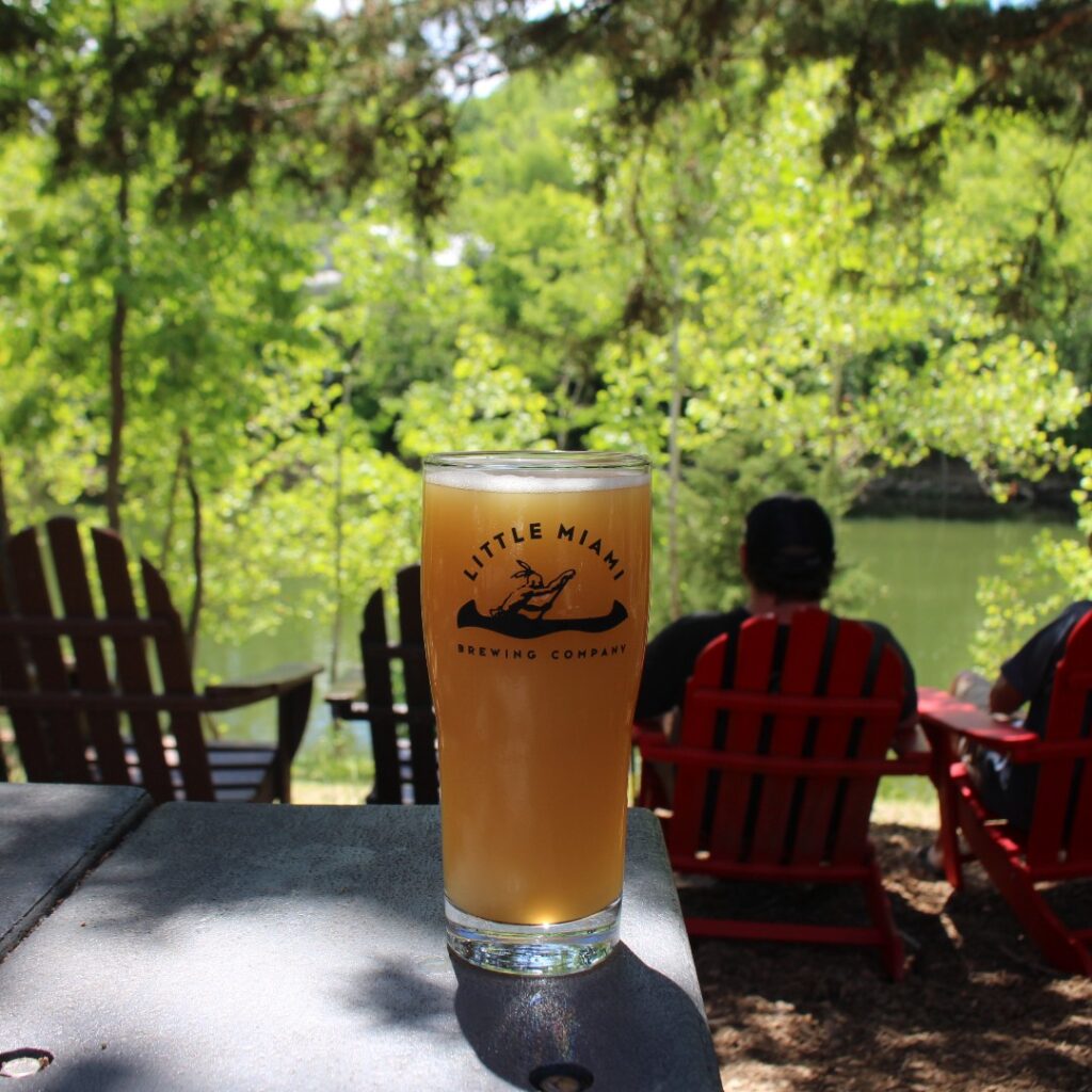 A pint of Little Miami Brewing Company beer on a table overlooking the river with guests relaxing nearby.