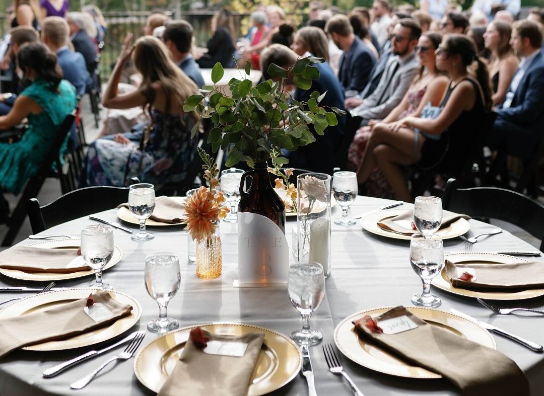 An elegant outdoor reception table setting at Little Miami Brewing Co., with wedding guests seated in the background.