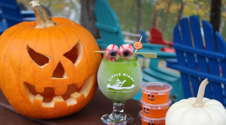 A spooky riverside setup with a jack-o'-lantern, green cocktail, jello shots, and a flyer for a Halloween bash at LMBC.