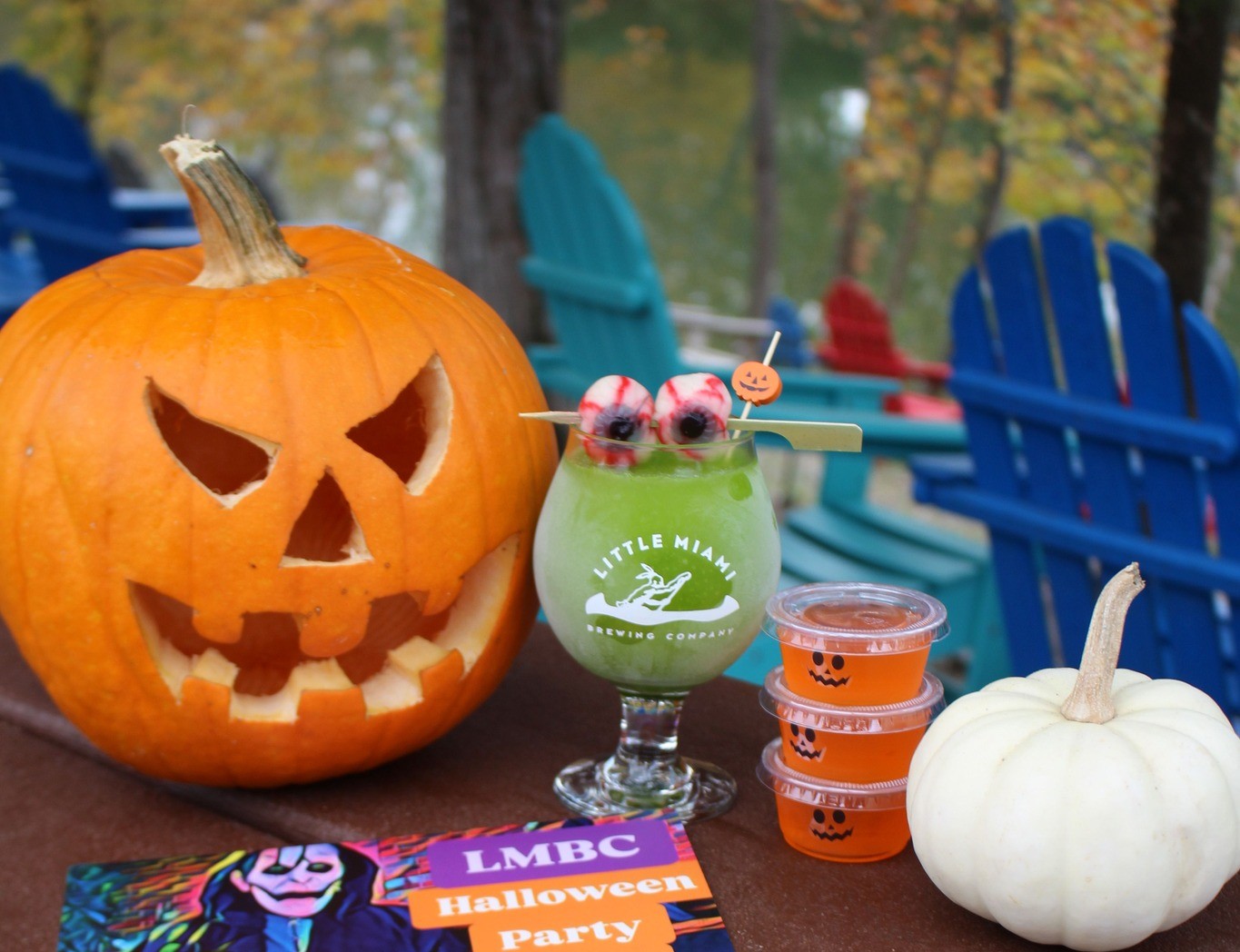 A spooky riverside setup with a jack-o'-lantern, green cocktail, jello shots, and a flyer for a Halloween bash at LMBC.