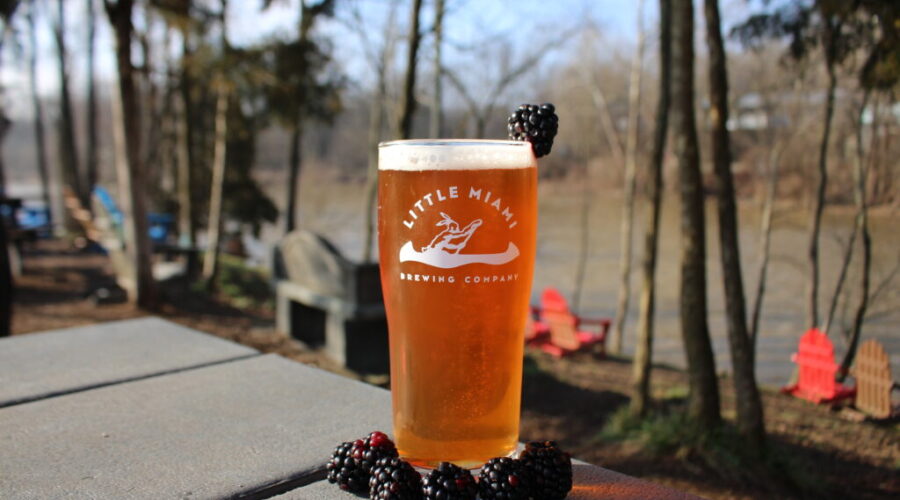 Little Miami Brewing Company’s Blackbird Fly blackberry wheat beer on a table with fresh blackberries and the Little Miami River in the background.
