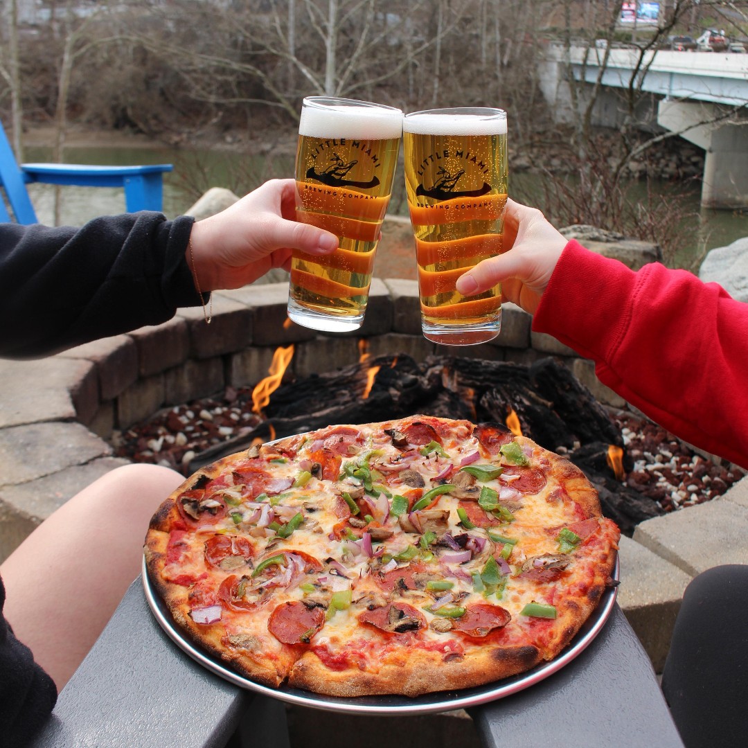 A freshly baked pizza paired with clinking beer glasses beside the fire pit at Little Miami Brewing Company.