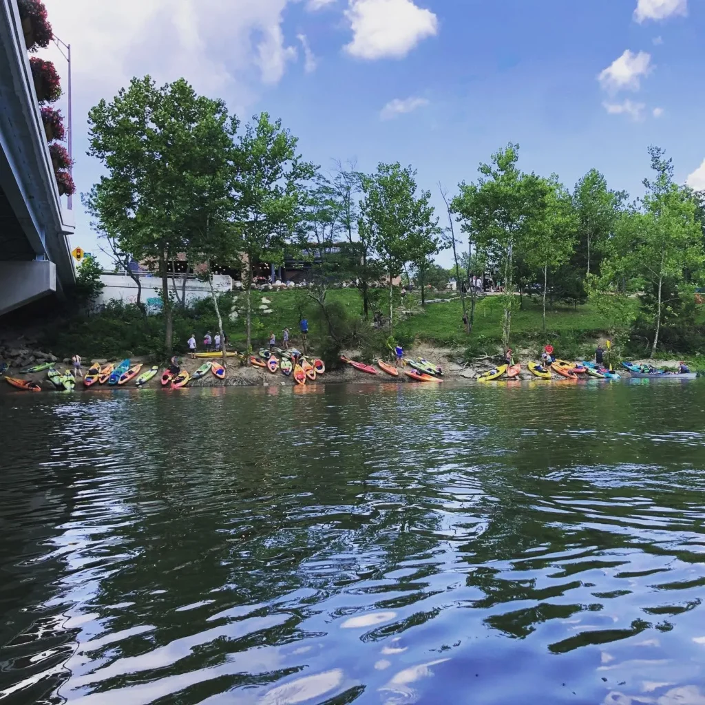 Colorful kayaks and gear lined up on Little Miami riverbank ready for rental beside Little Miami Brewing Company.