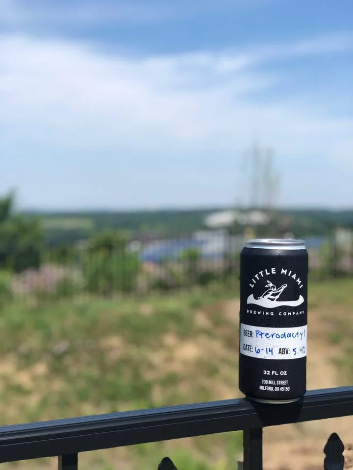 Labeled crowler can from Little Miami Brewing Company on an outdoor railing overlooking a scenic landscape.