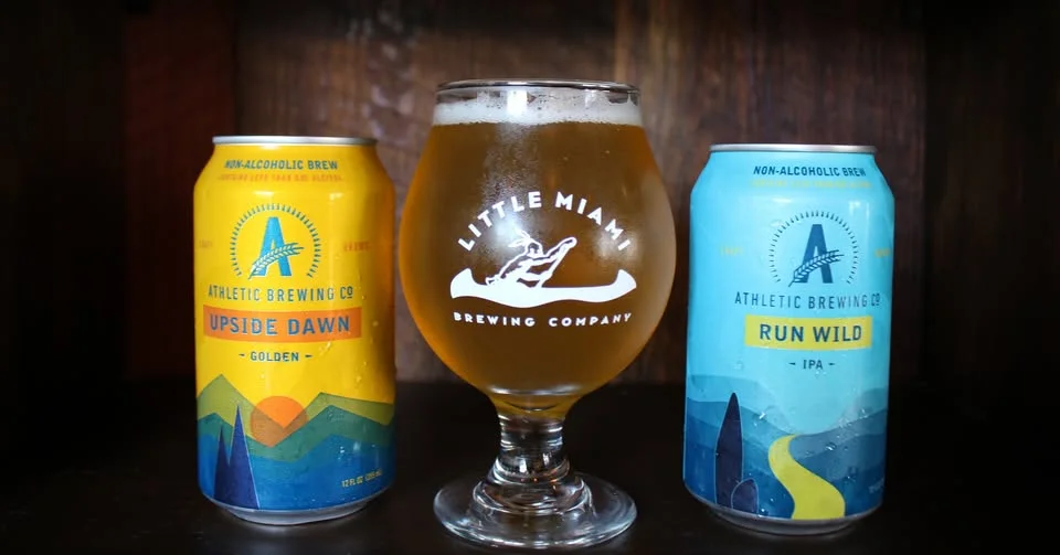 Two cans of non-alcoholic craft beer flanking a branded glass filled with a golden brew at Little Miami Brewing Company.
