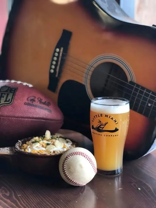 Leather rugby ball, baseball, acoustic guitar, and a craft beer arranged for game day at Little Miami Brewing Company.