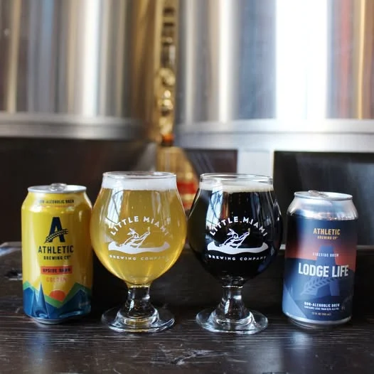 Two branded glasses of non-alcoholic beer displayed beside cans of golden and dark brews at Little Miami Brewing Co.